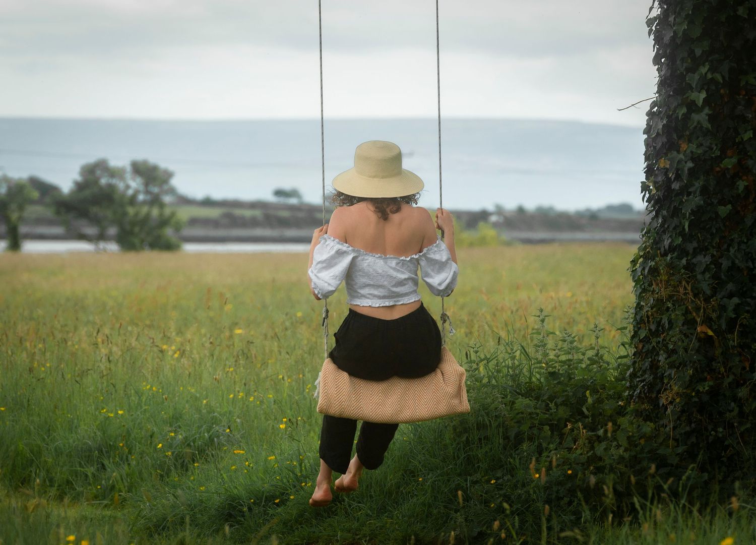 Woman in hat sitting on rope swing facing ocean in green meadow with yellow wildflowers.
