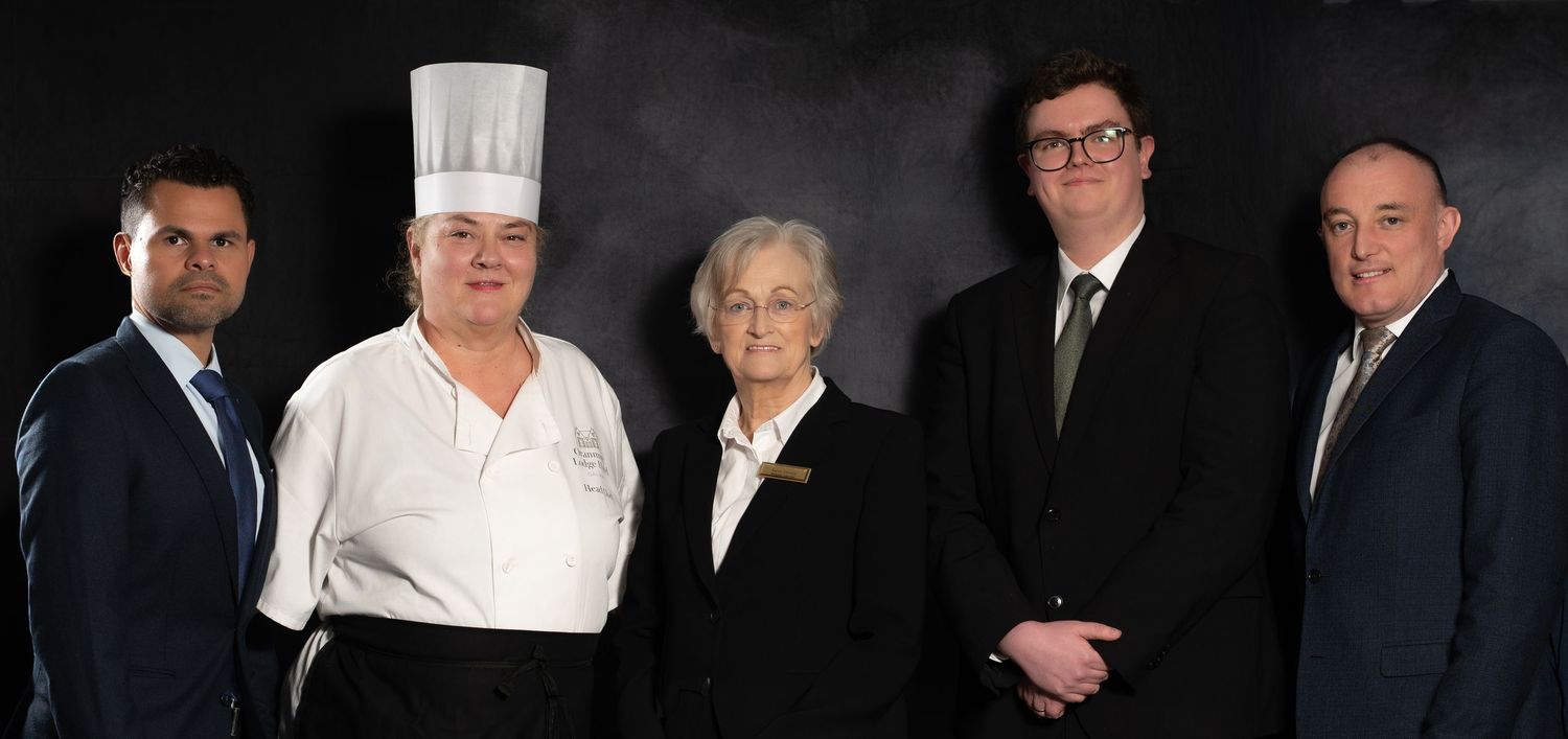 Five hospitality professionals posing formally, including a chef in white uniform and others in business attire, against dark background.
