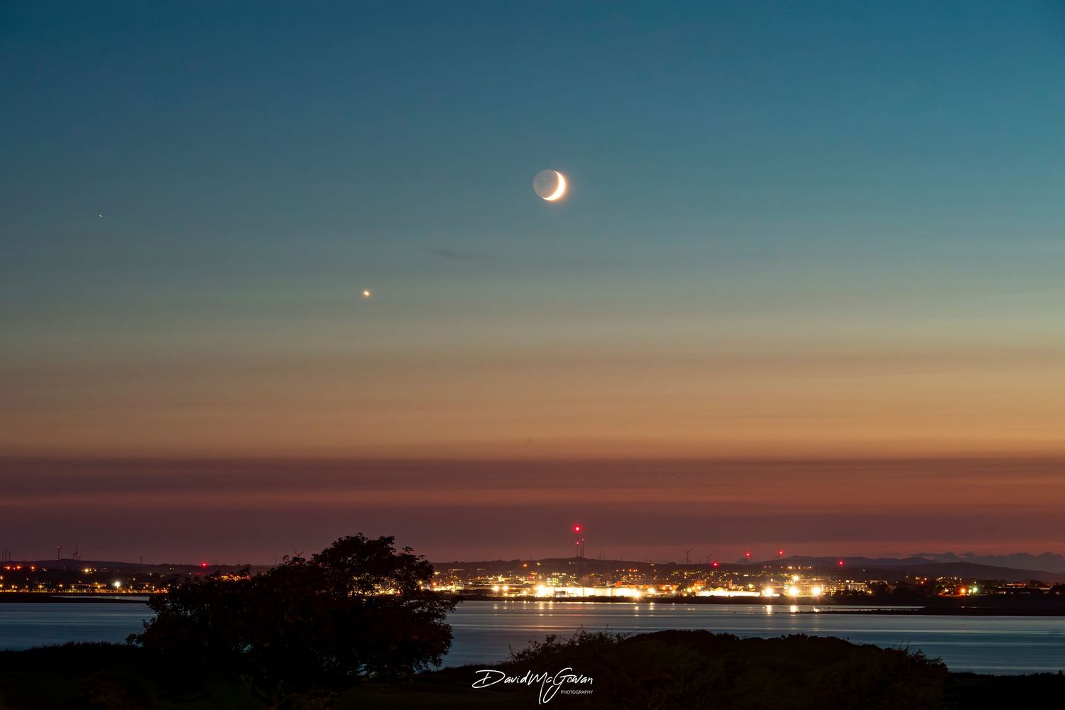 Crescent moon and planet Venus shine over a coastal city at twilight with colorful sunset hues.