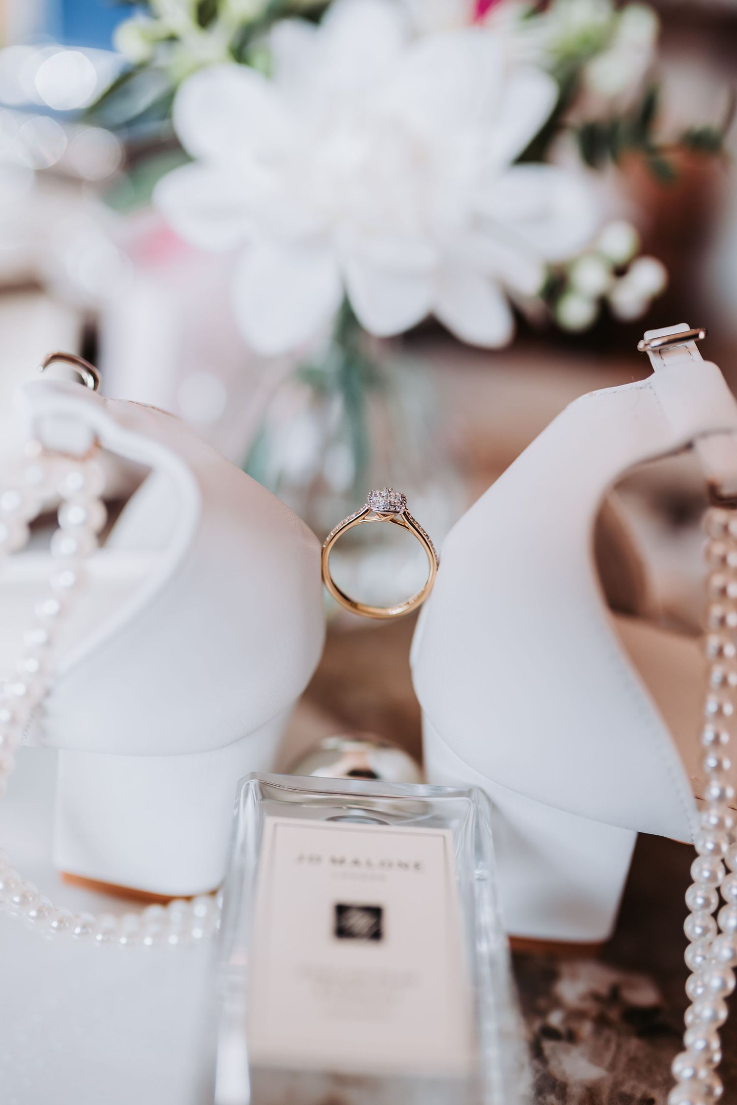 Wedding details including rings, pearls, and Jo Malone perfume arranged on white surface with soft lighting.