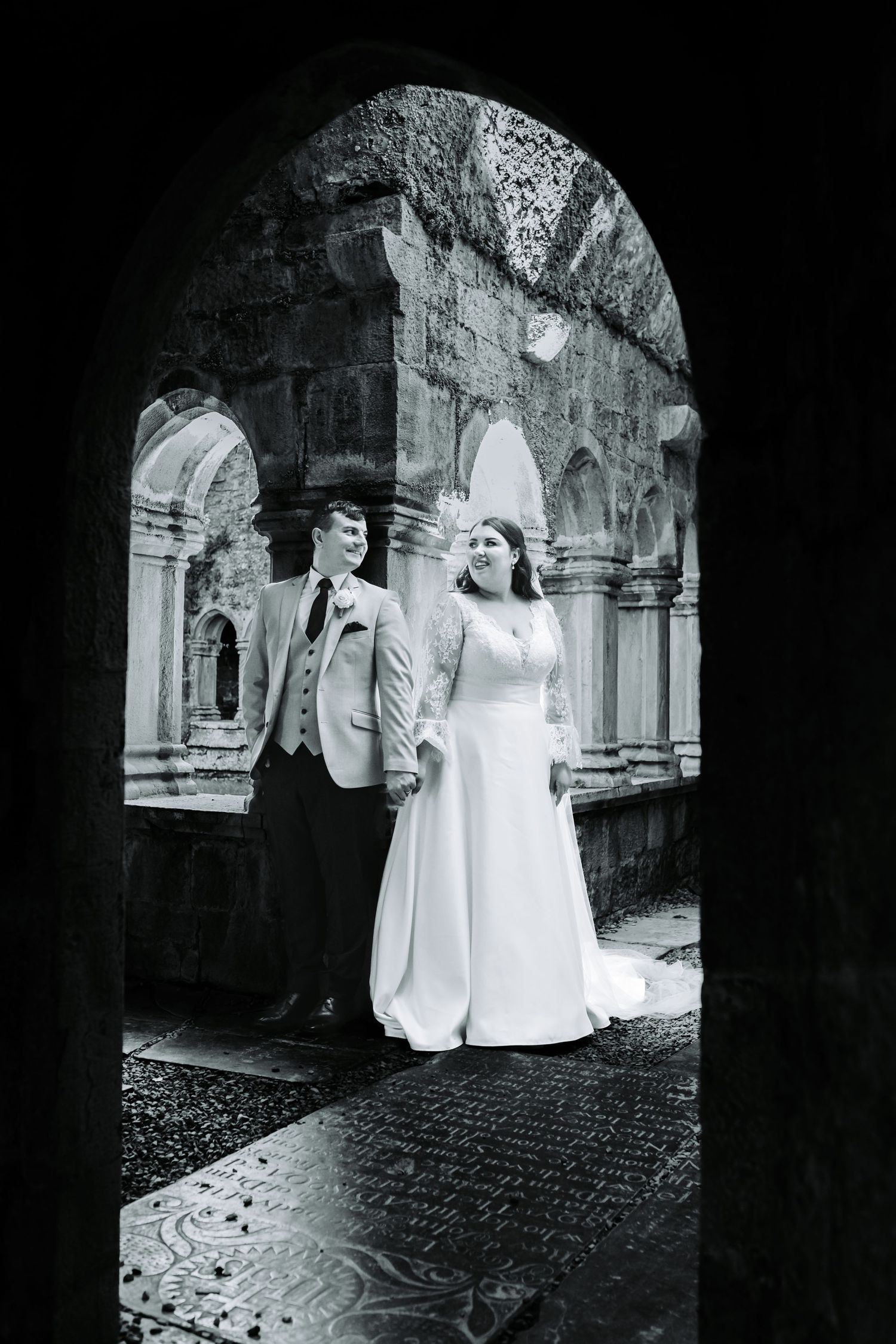 Black and white photo of bride and groom holding hands under a historic stone abbey archway.