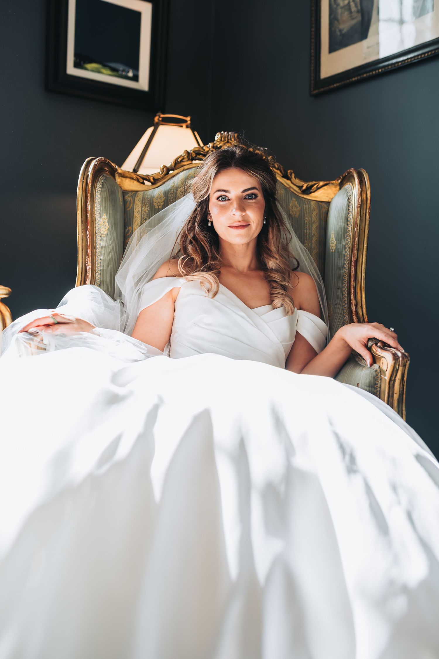 A beautiful bride in a white off-shoulder gown sits elegantly in a gold ornate armchair.