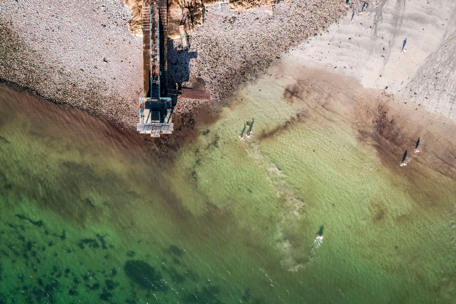 Aerial top-down view of a pier and swimmers in clear green coastal waters.