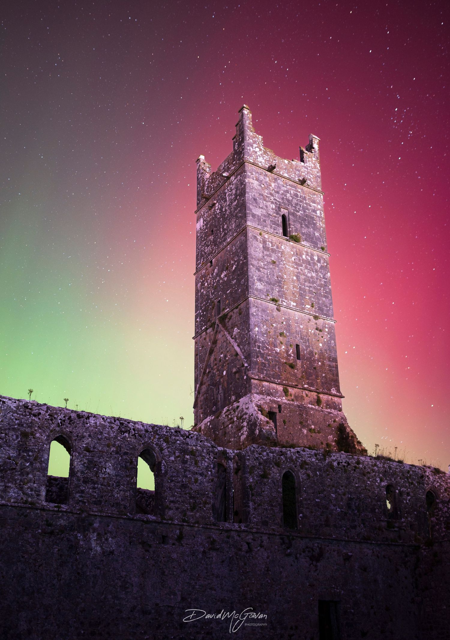 A medieval stone castle tower illuminated by vibrant green and red northern lights in a starry sky.