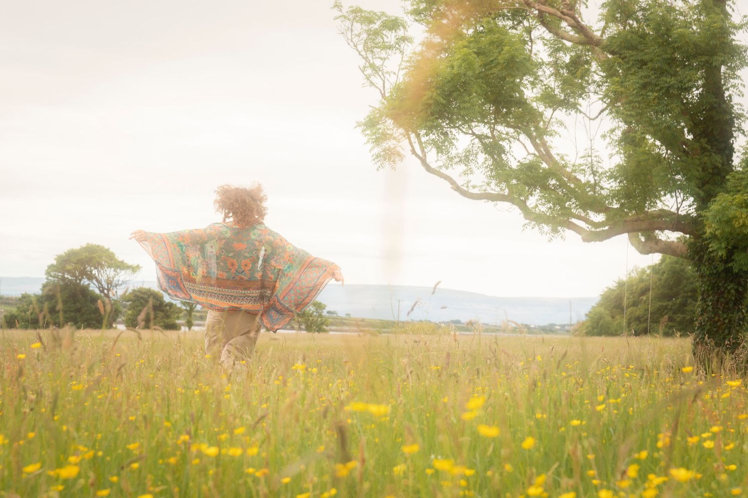 Woman in colorful kimono running through a sunny wildflower meadow with yellow blooms and green trees.