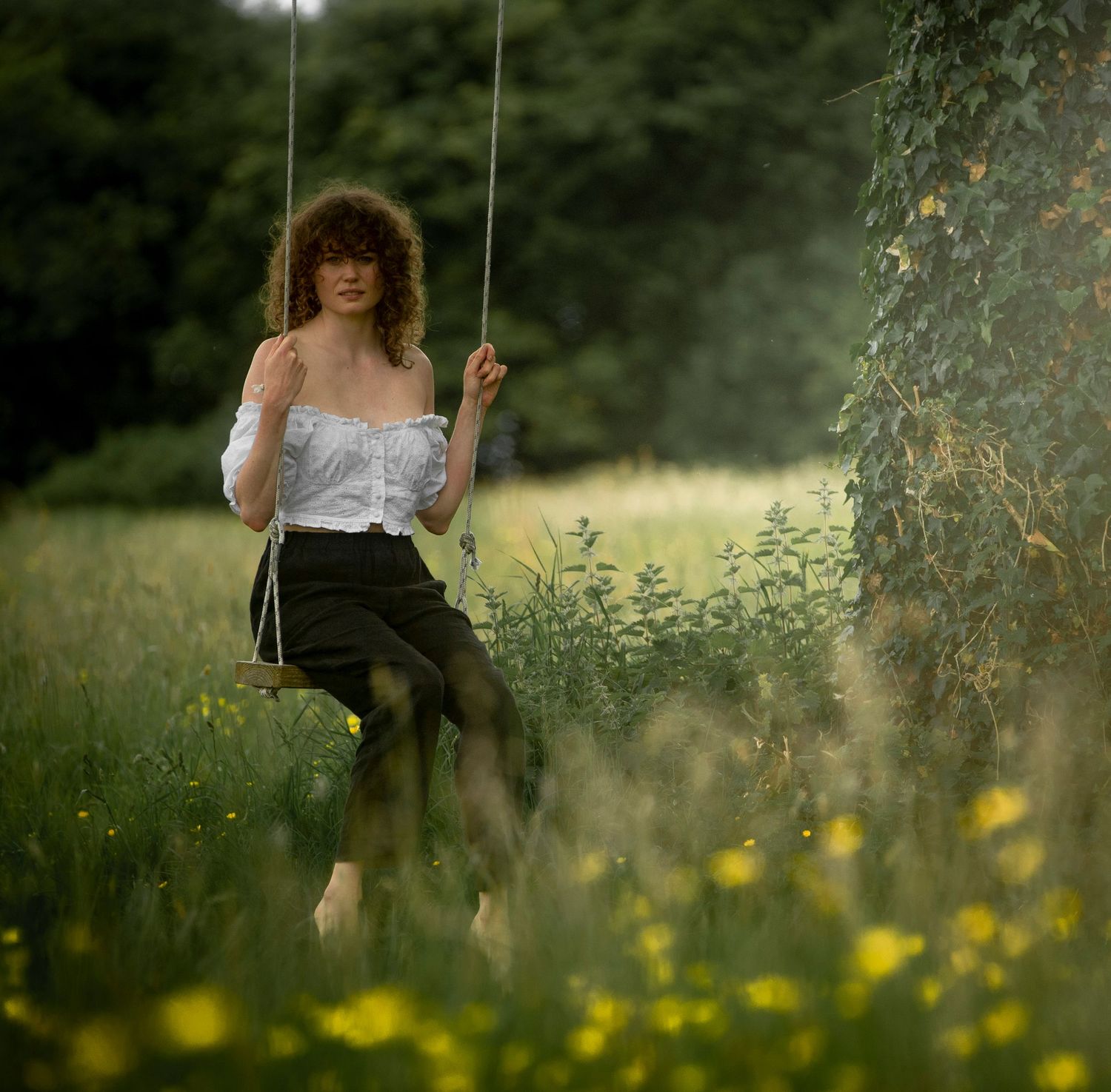 Curly-haired woman sitting on rope swing in lush green meadow with yellow wildflowers.