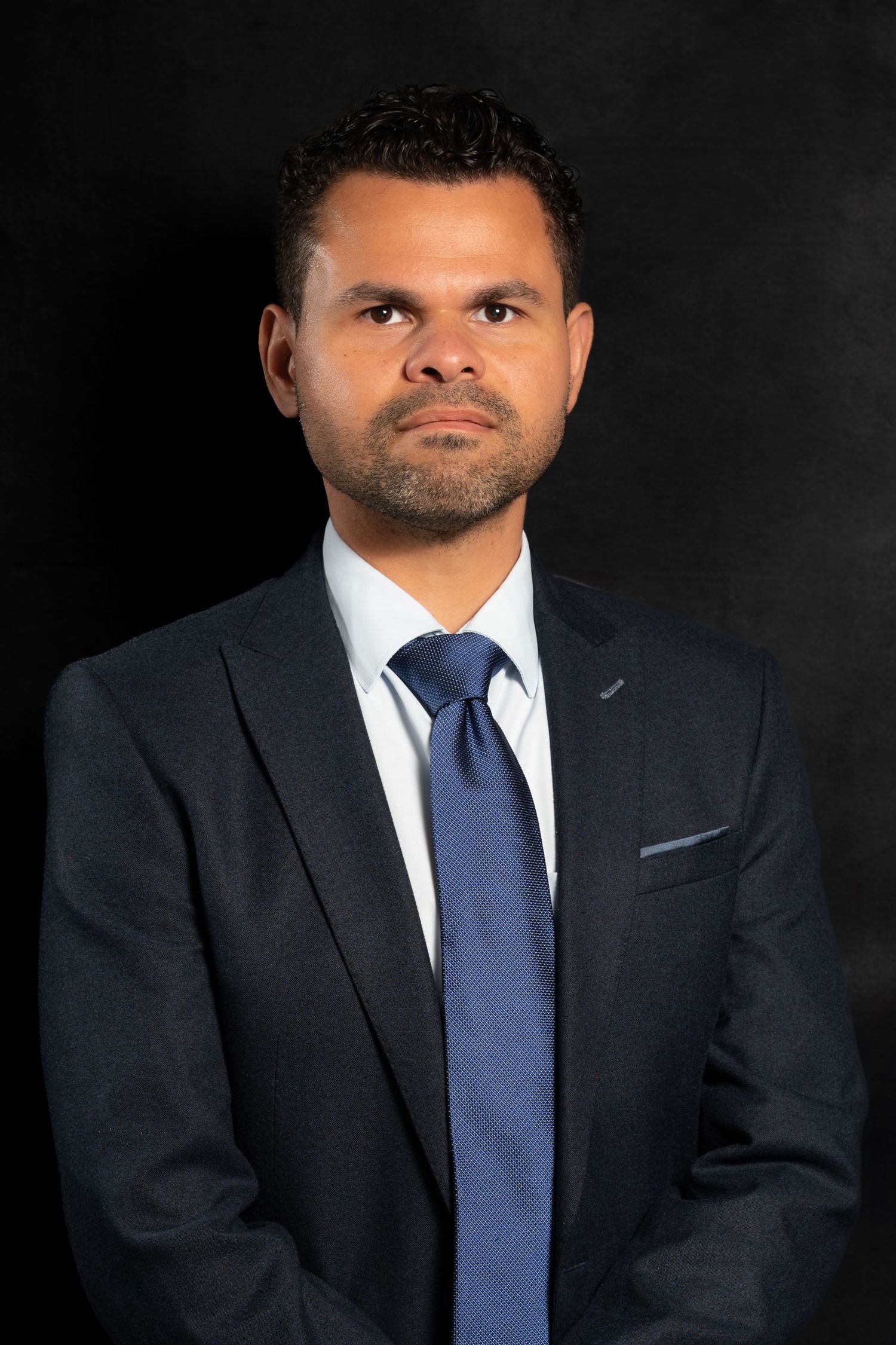 Professional man in dark suit and blue tie poses for formal corporate headshot photo.