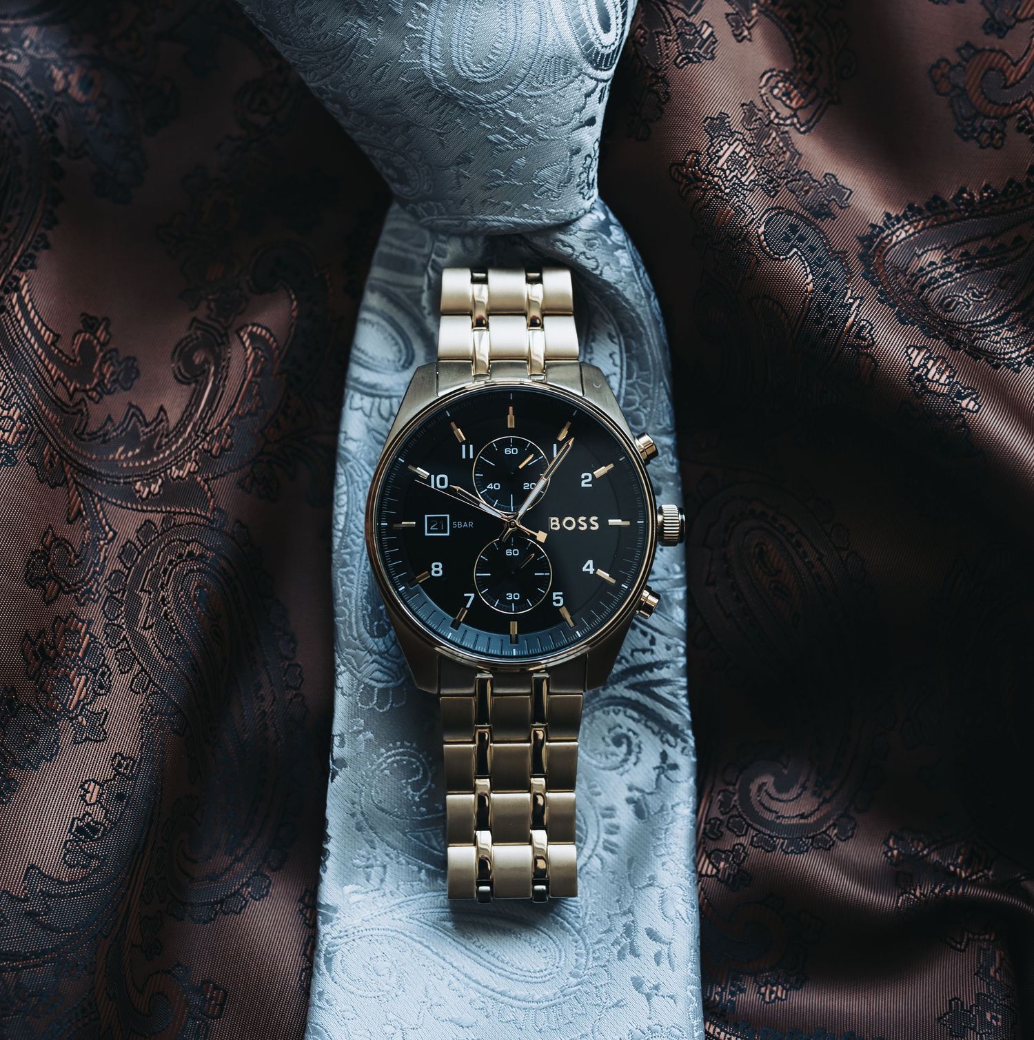 A silver chronograph watch rests on textured fabric with elegant patterns.