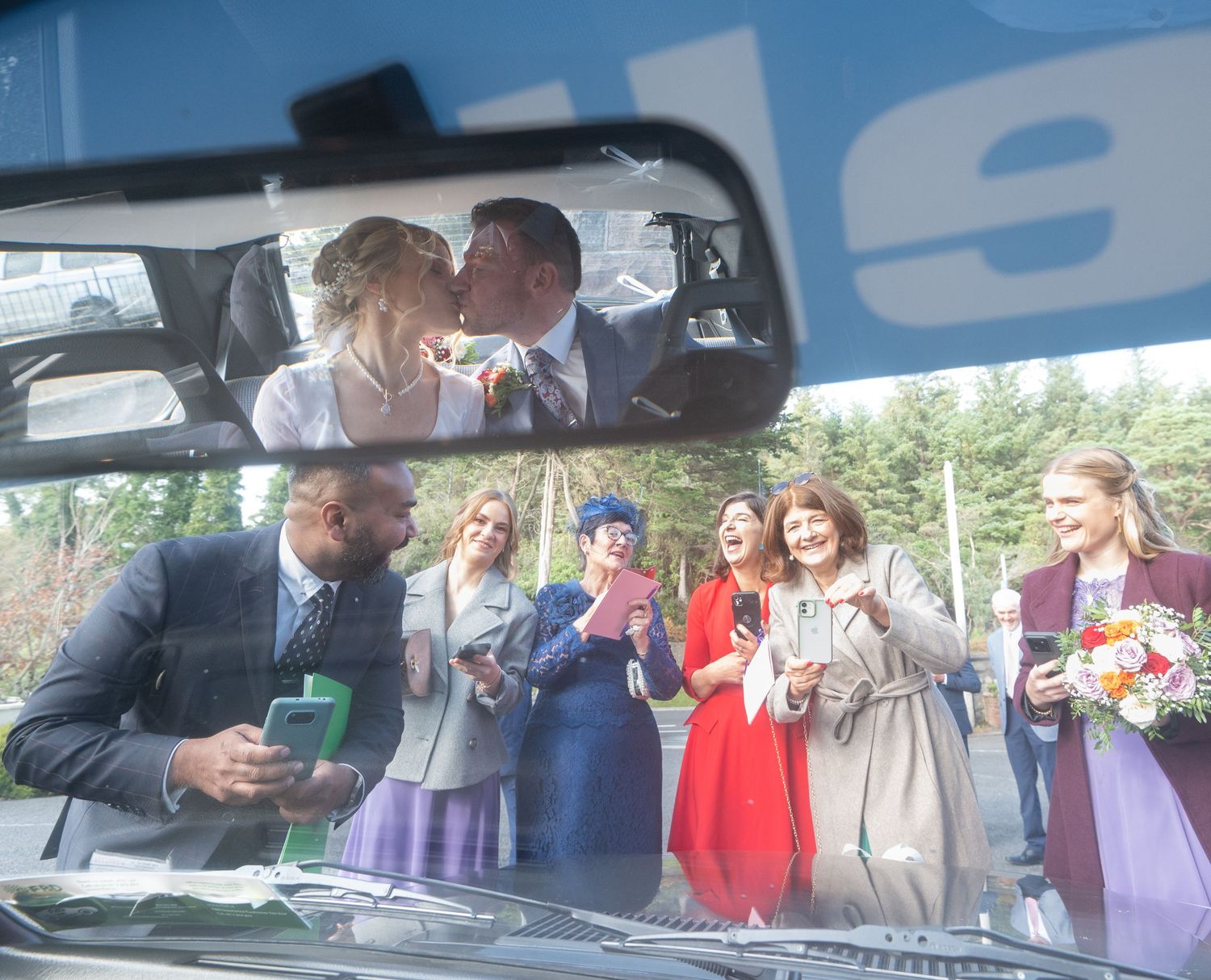 Wedding guests gather around a car at an outdoor reception celebration.