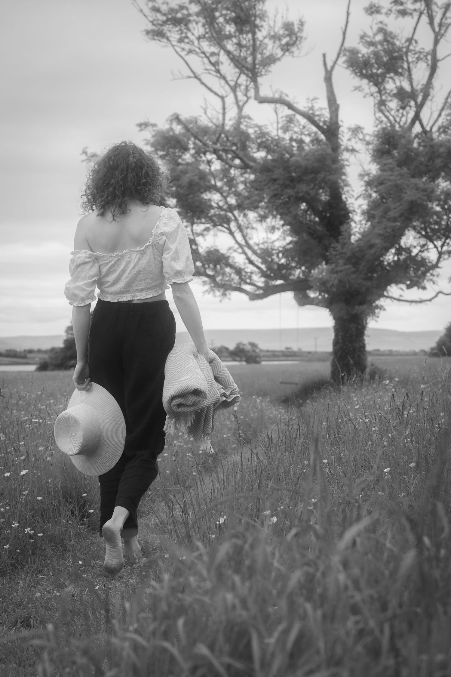 Woman with curly hair walking barefoot in field carrying hat, black and white.