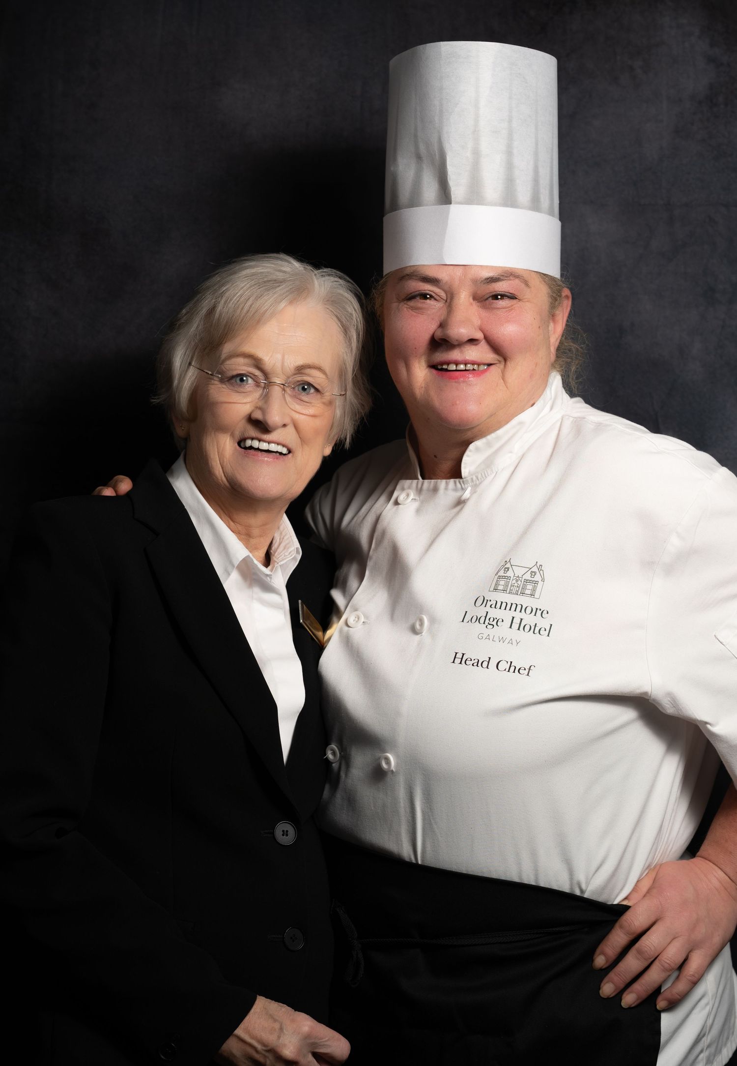 Elderly woman poses with head chef in white uniform and tall hat for professional portrait.