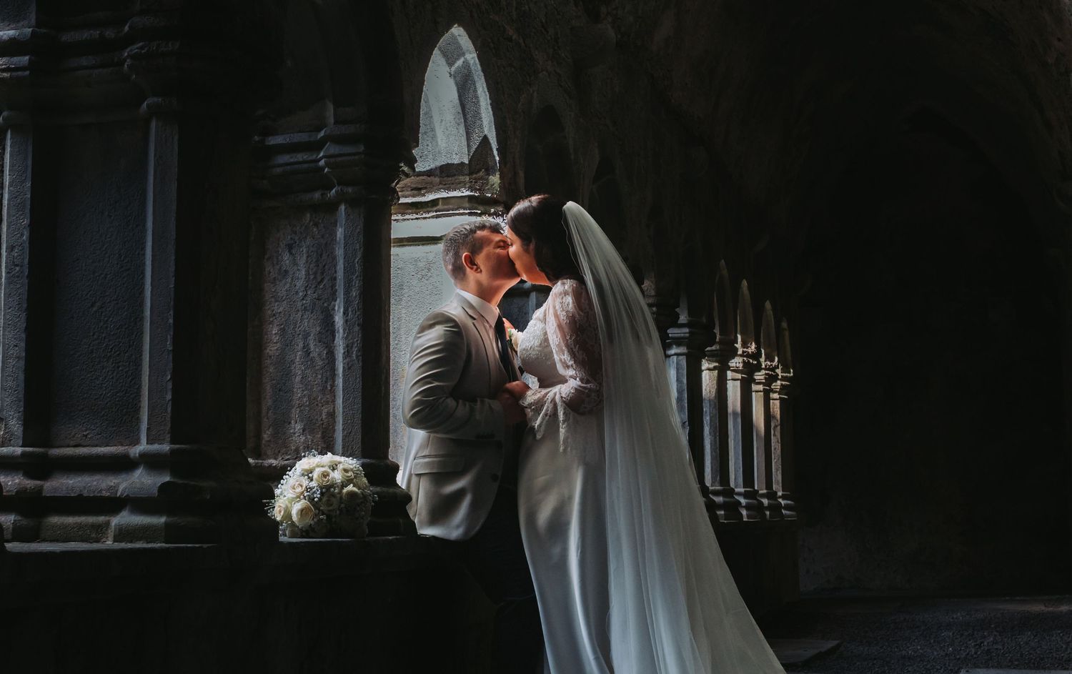Bride and groom sharing a romantic kiss inside a dramatic dark stone church archway.