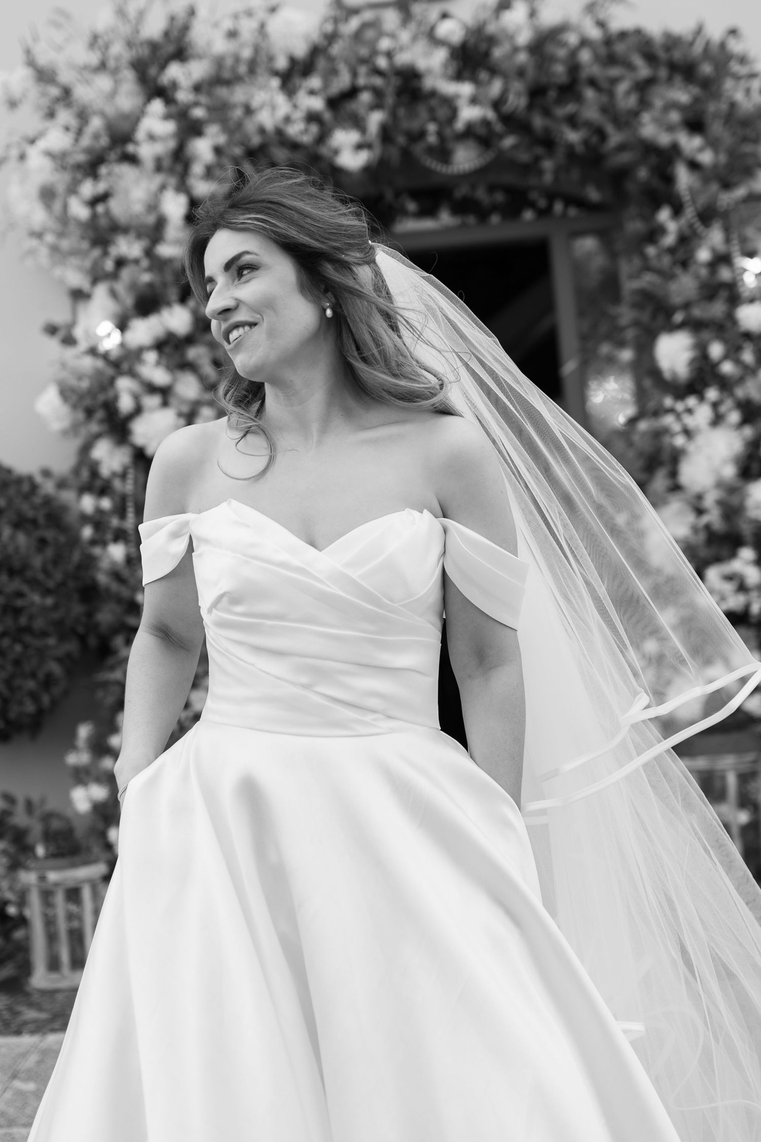Black and white portrait of bride in off-shoulder gown and veil outdoors at wedding venue.