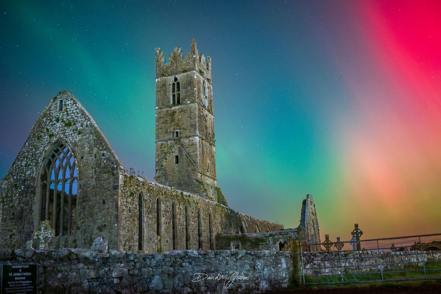 Ancient Irish stone abbey ruins illuminated by vibrant aurora borealis colors in night sky.