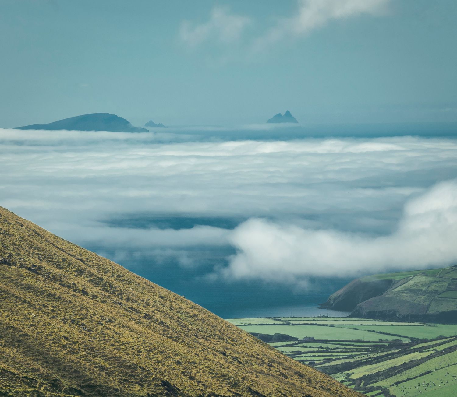 Islands emerge above a sea of clouds with green coastal fields visible on the Dingle Peninsula, Ireland.