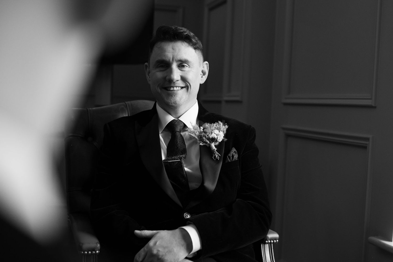Black and white photo of a smiling groom in a suit with boutonniere seated at a wedding ceremony.