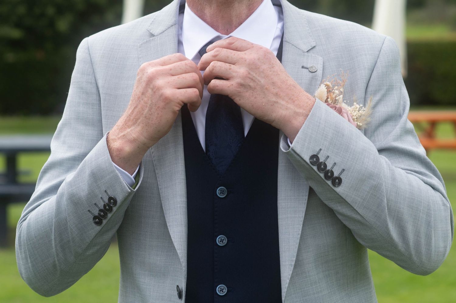 Man in grey suit adjusting navy tie with floral pocket square outdoors.