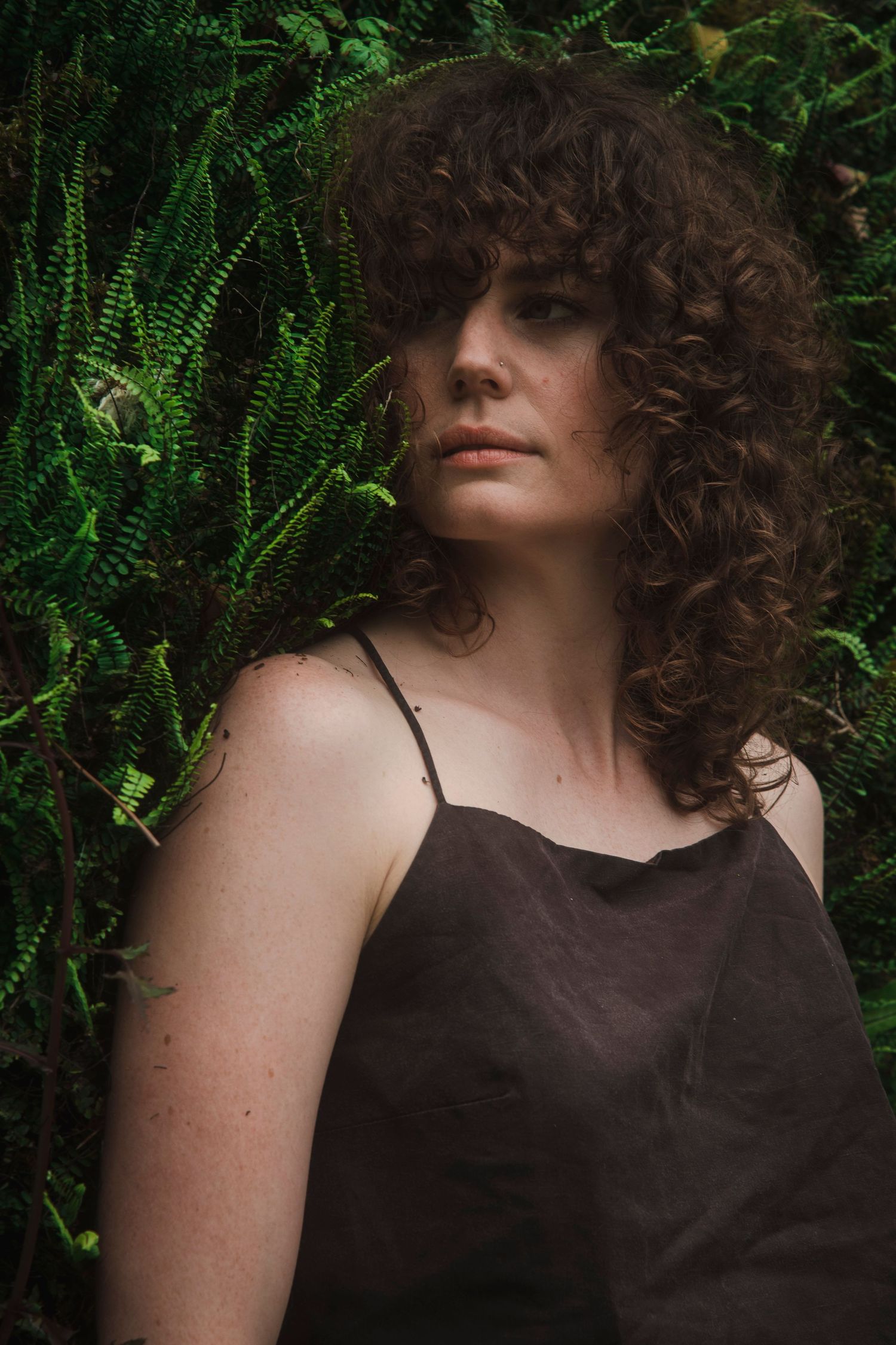 Woman with curly dark hair in brown camisole posing against lush green ferns.