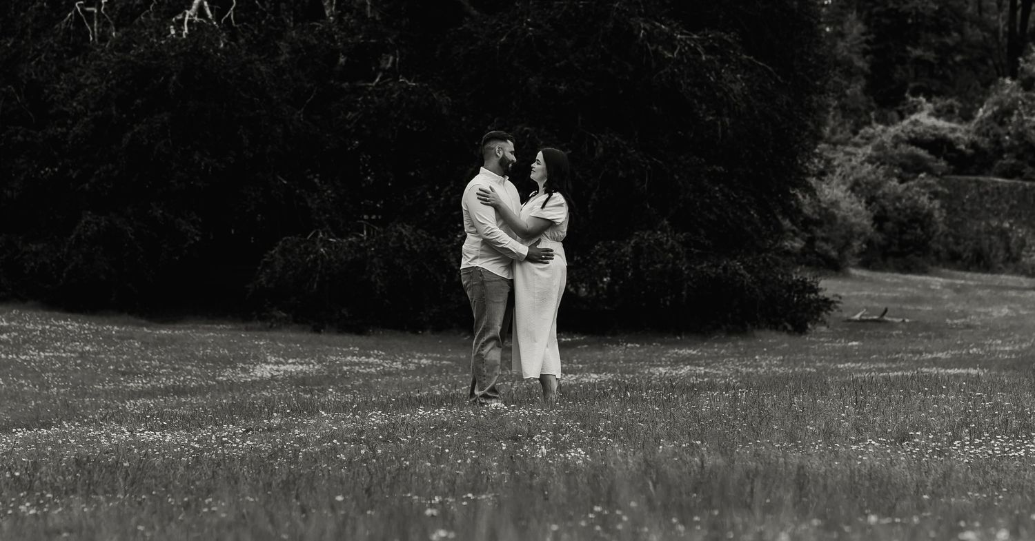 Black and white photo of a couple embracing in a lush green meadow surrounded by trees.
