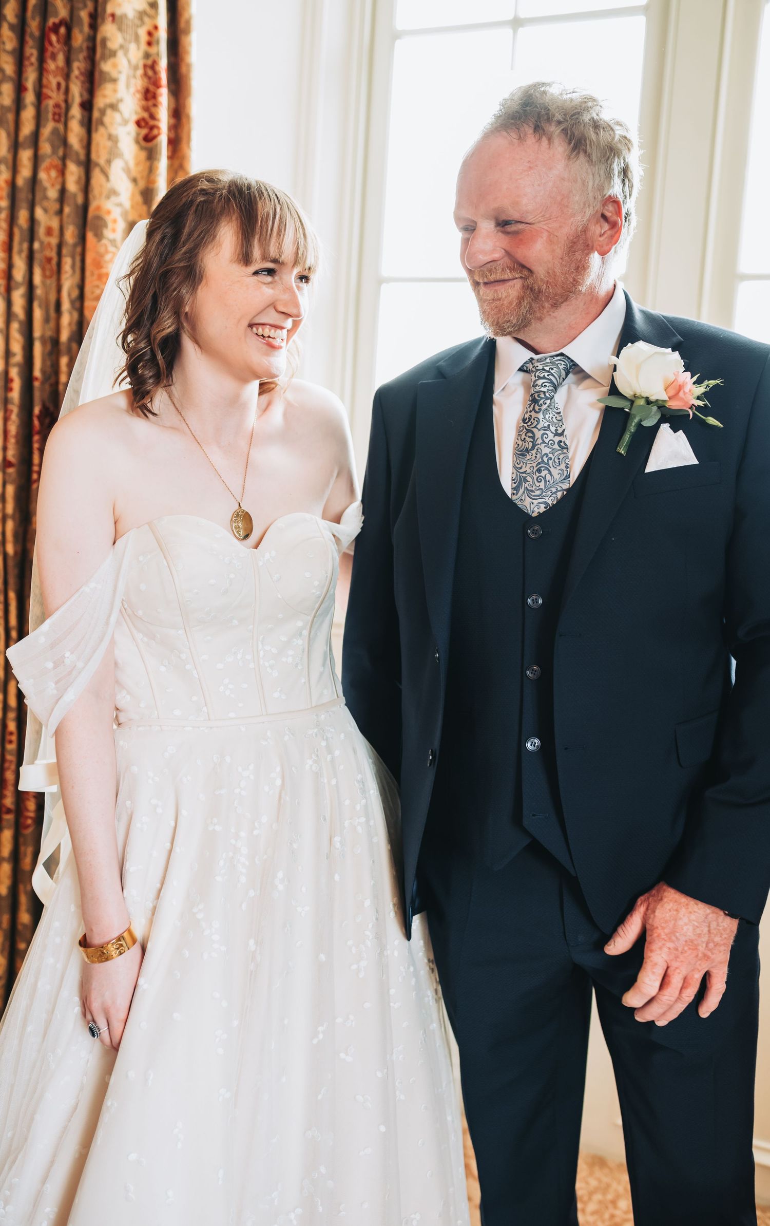 Smiling bride in white gown and veil stands with older man in navy suit with floral boutonniere at wedding.