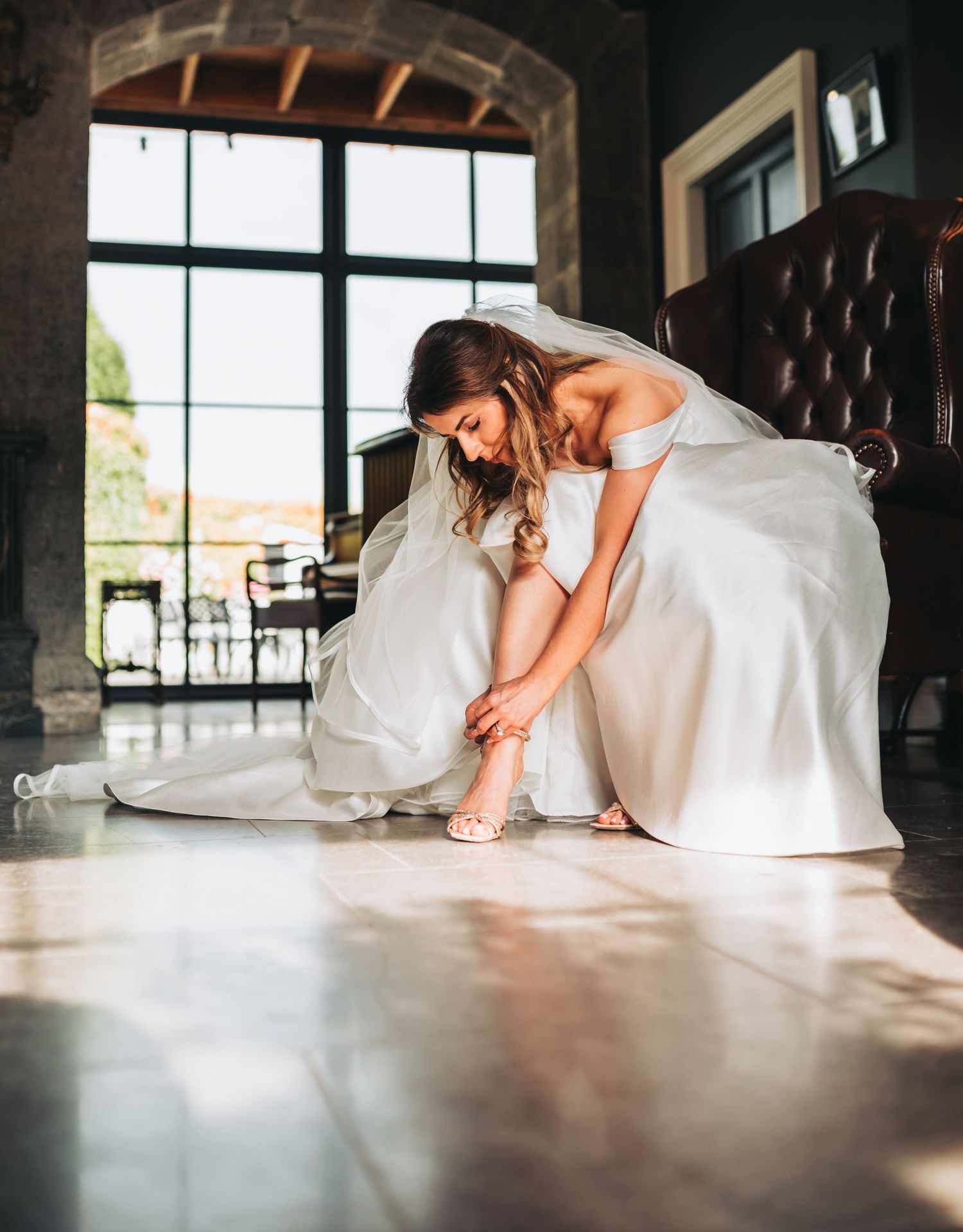 A bride in a flowing white gown adjusts her shoes in a rustic venue with large windows.