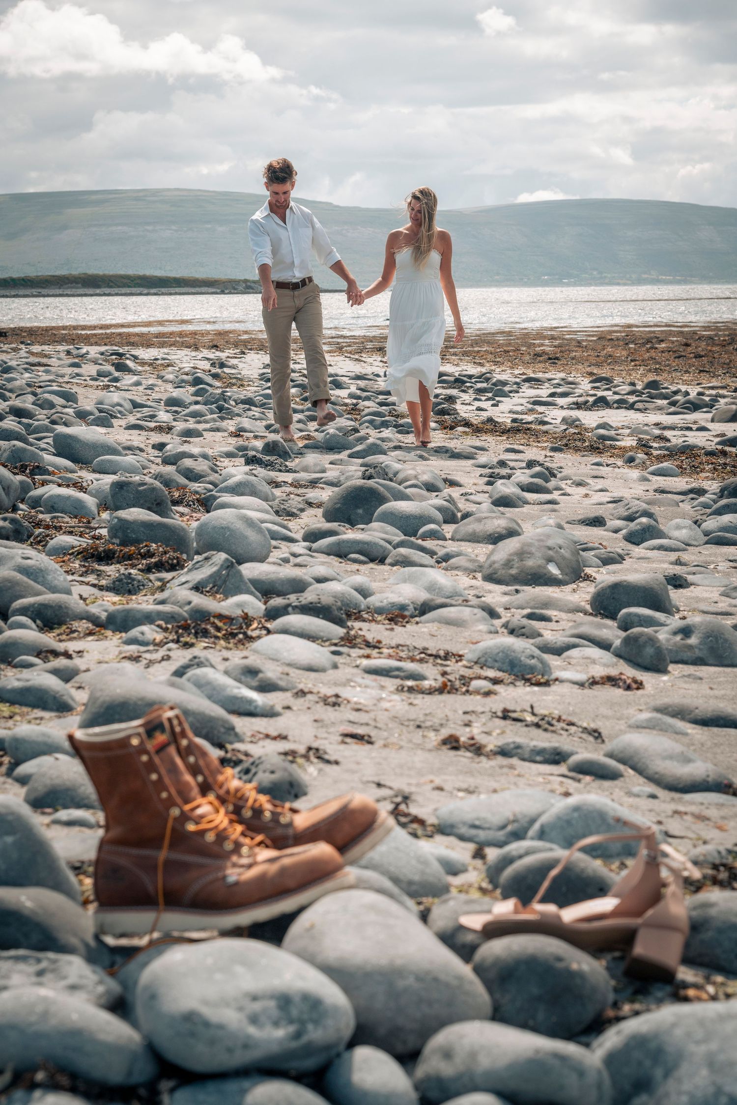A couple walks along a rocky beach with boots in the foreground and mountains in the background.