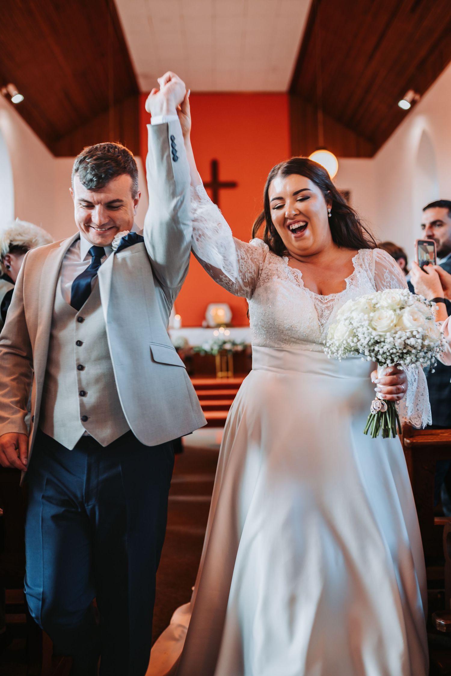 Joyful newlyweds raising hands walking down church aisle after wedding ceremony.