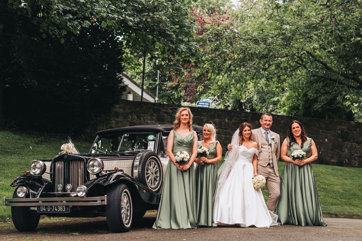 Bride with bridesmaids in sage green gowns posing beside a vintage classic wedding car.