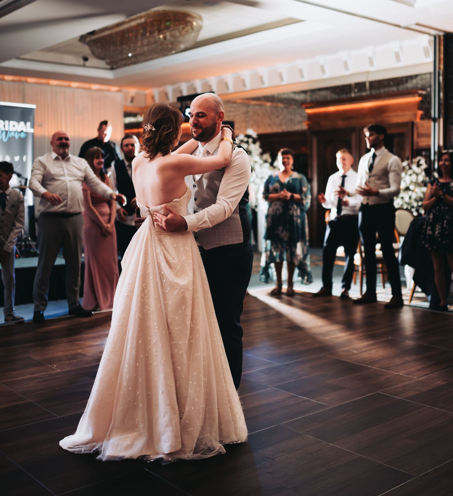 Bride and groom sharing romantic first dance in ballroom while wedding guests watch in background.
