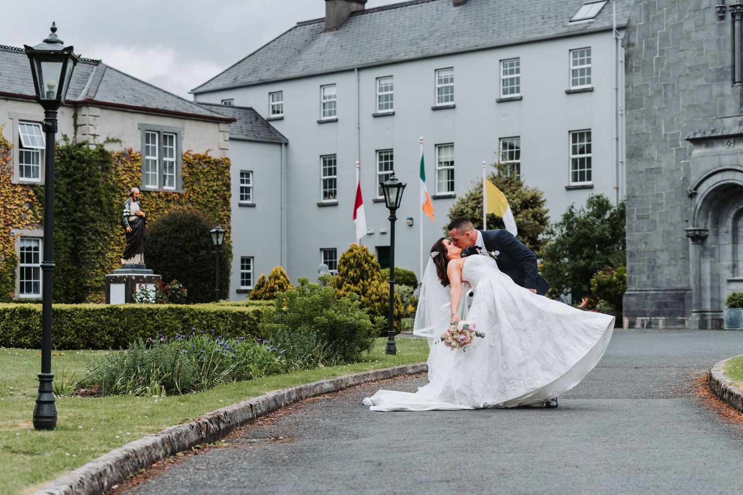 Bride and groom share a romantic dip kiss on a curved driveway outside an elegant white Irish manor wedding venue.