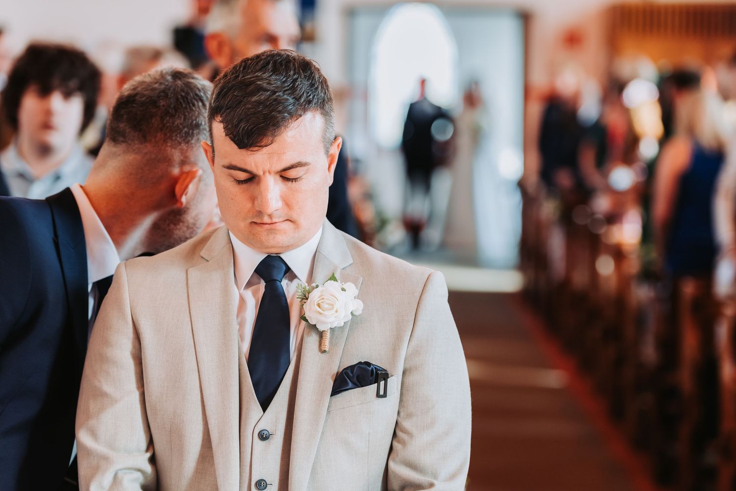 Groom in beige suit with white boutonniere standing solemnly at church altar before ceremony.