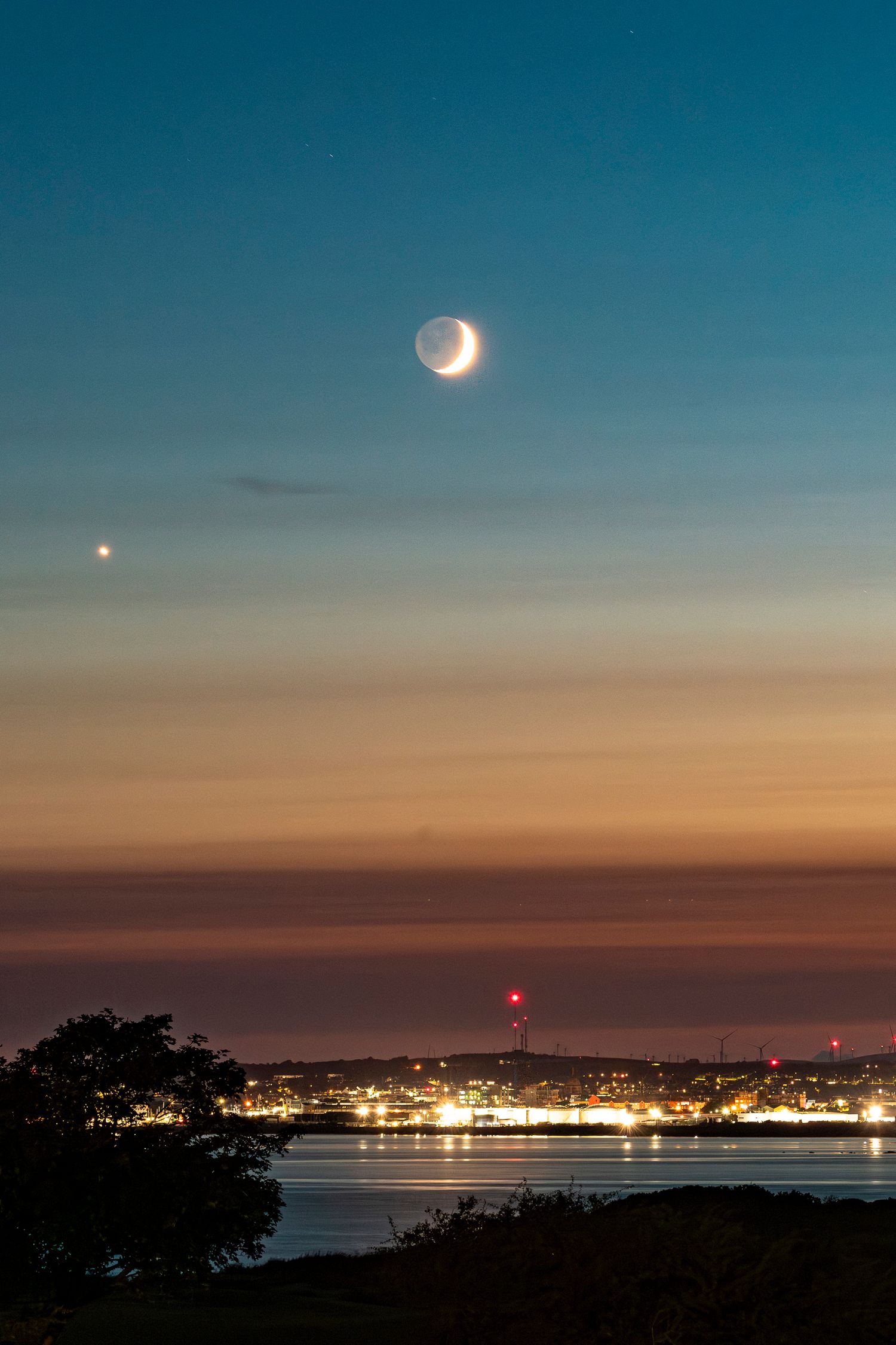 Crescent moon and Venus shine over a coastal city at twilight with warm orange and blue sky tones.
