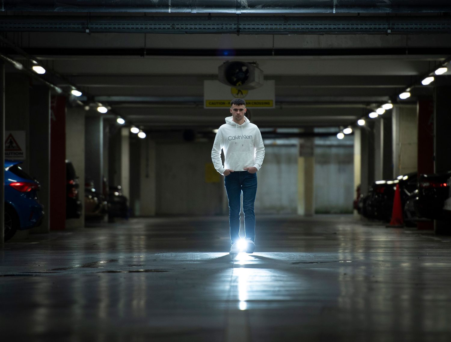 Man in white hoodie walking through a dimly lit underground parking garage with bright light glowing behind him.