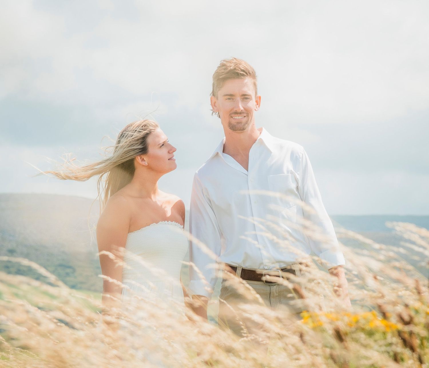 A romantic couple shares a joyful moment together on a windswept hilltop overlooking a scenic landscape.