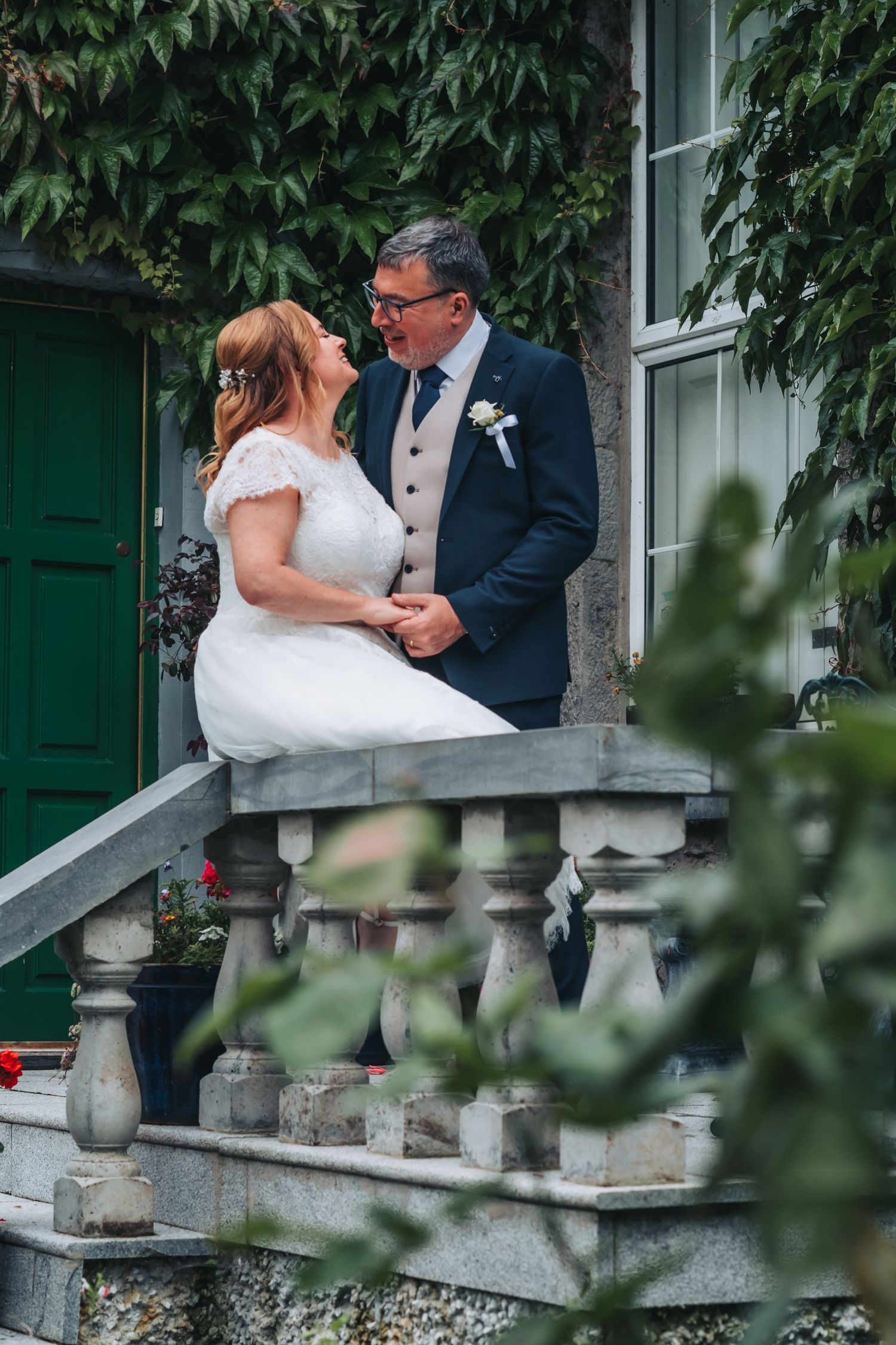 Newlyweds share a romantic moment on an elegant stone balcony surrounded by greenery.