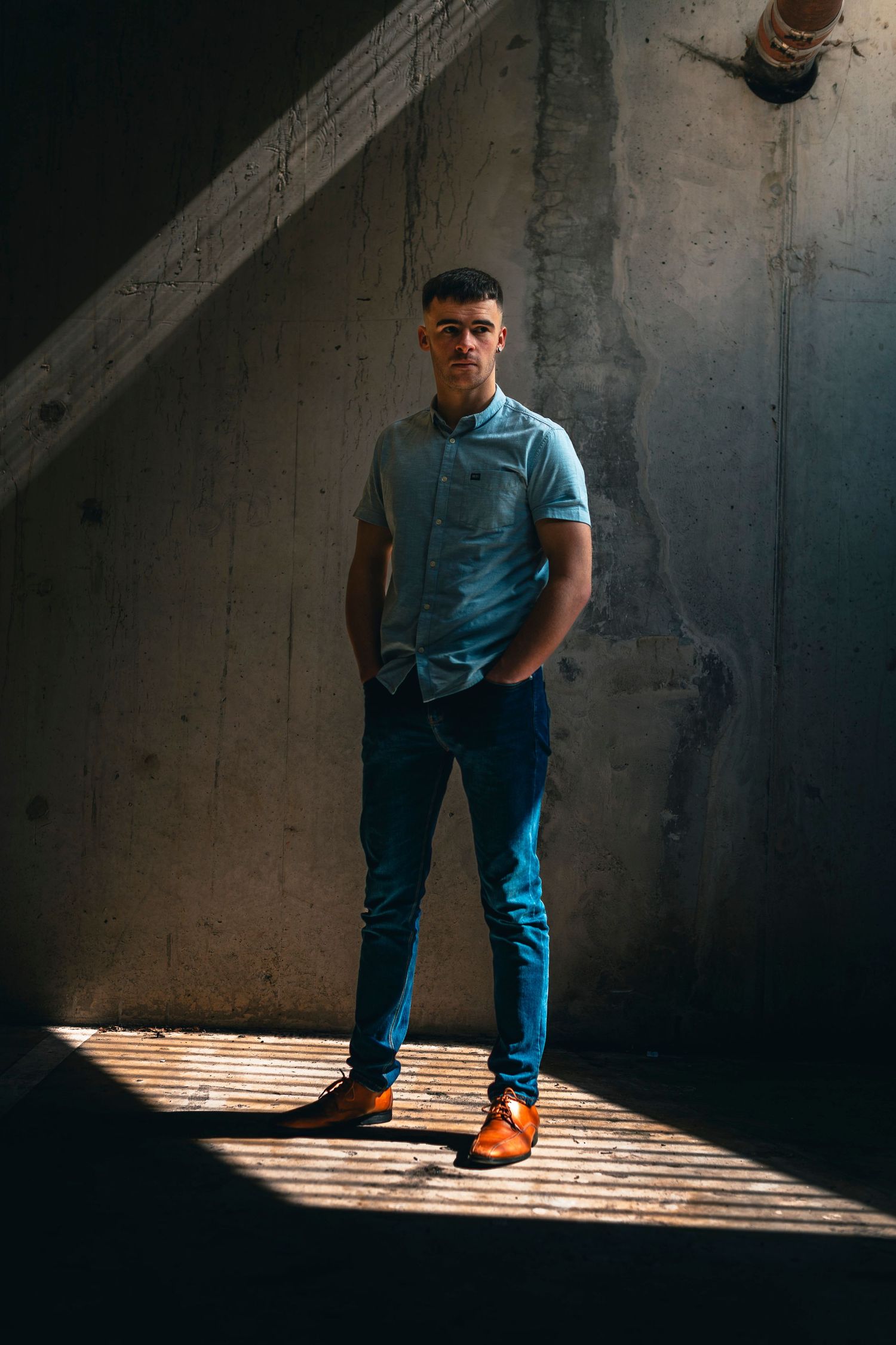 Young man in blue polo shirt and jeans poses confidently in dramatic shaft of light against concrete wall.