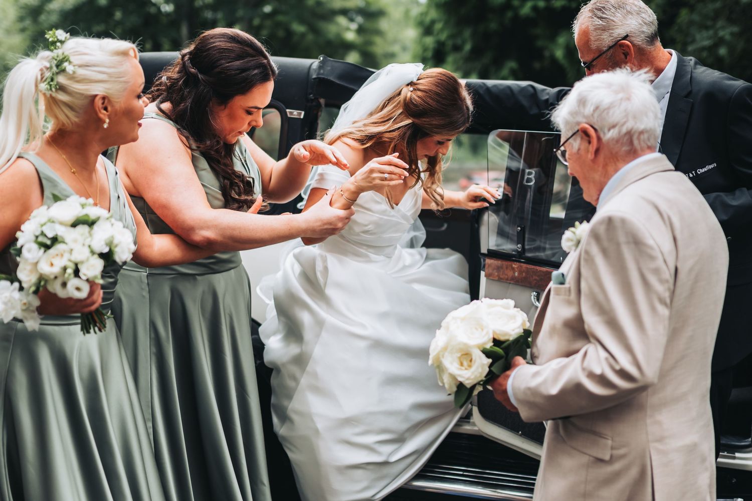 Bride in white gown assisted by bridesmaids in sage green dresses and elderly man exiting vintage black car.