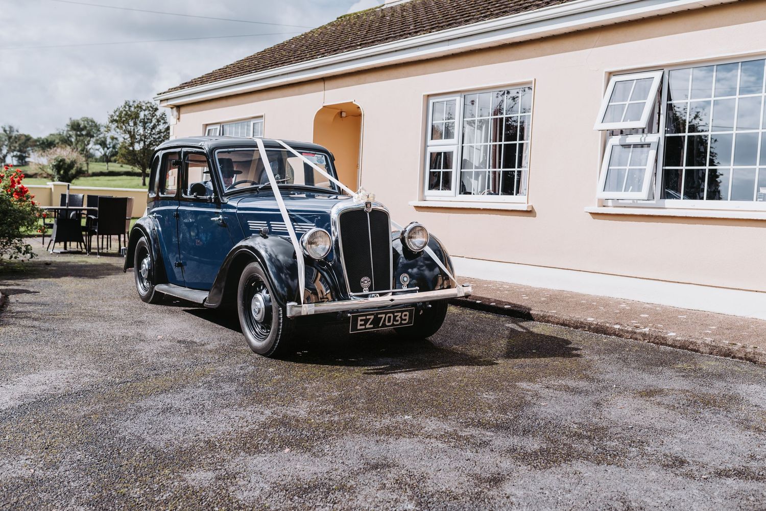 Vintage navy blue classic car decorated with white ribbon parked outside a wedding venue.