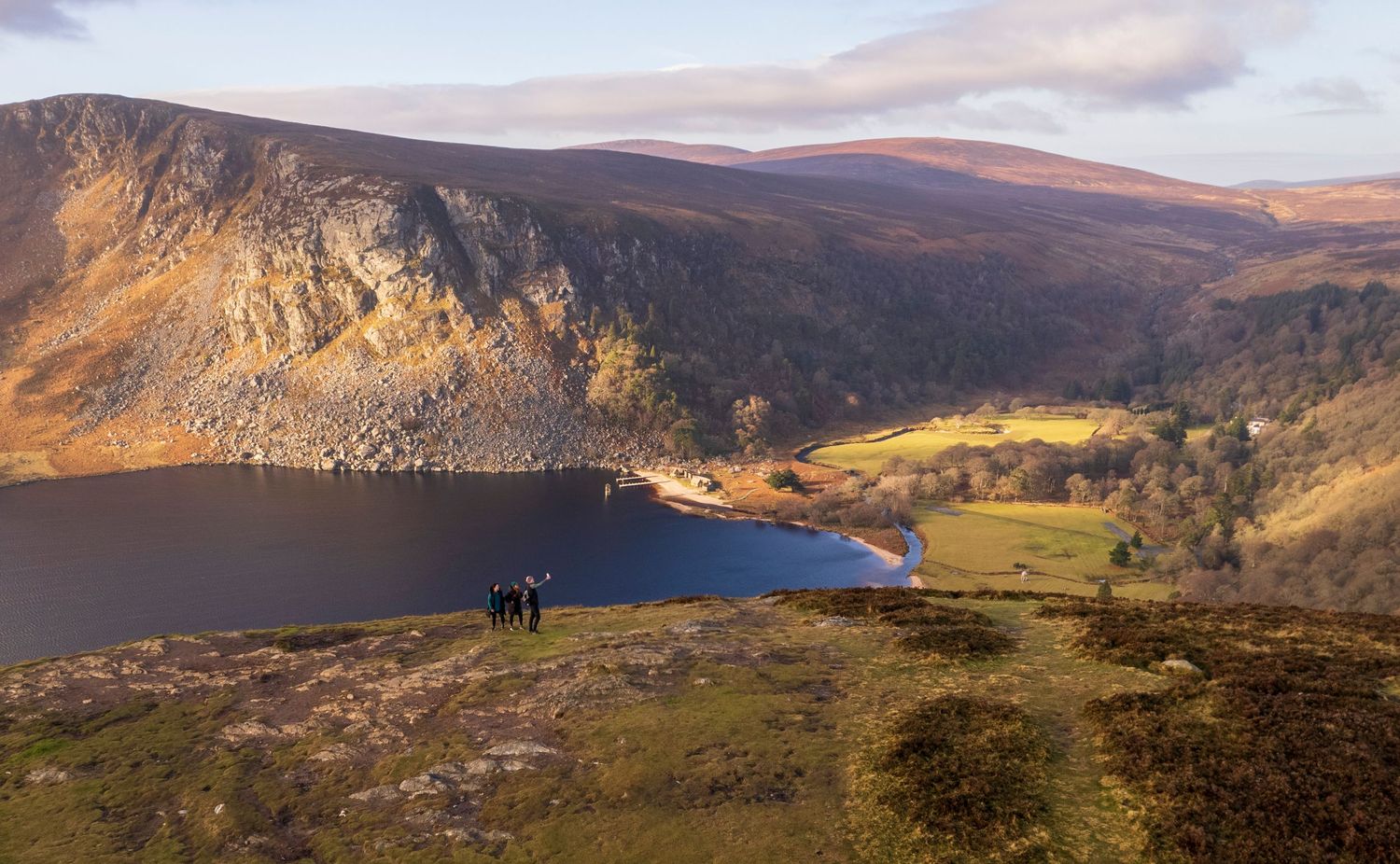 Hikers stand on a rocky summit overlooking a serene mountain lake surrounded by autumn hills in Ireland.