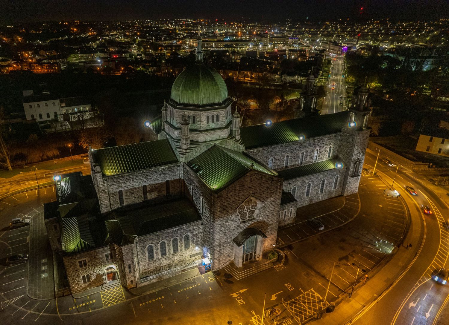 Aerial night view of Galway Cathedral illuminated against the city skyline with glowing streets below.