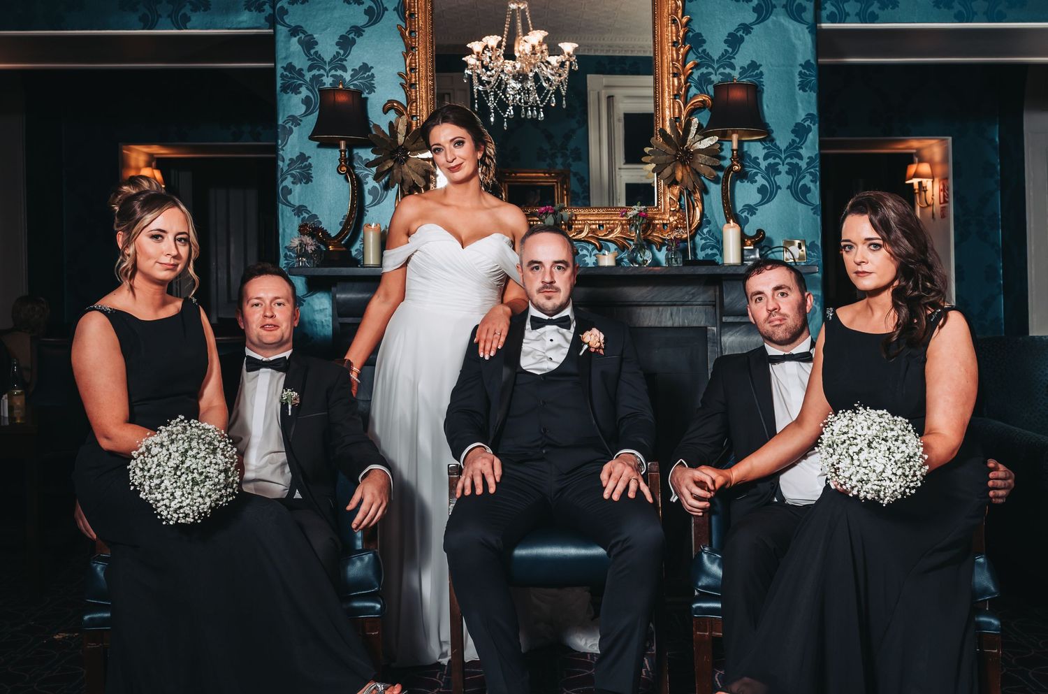 Wedding party posing elegantly in a blue Victorian room, bride in white gown with groomsmen and bridesmaids in black attire.
