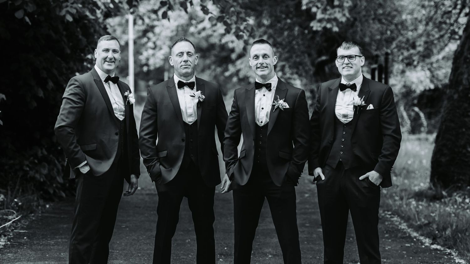 Four groomsmen in black tuxedos with bow ties and boutonnieres posing outdoors in a black and white wedding photo.