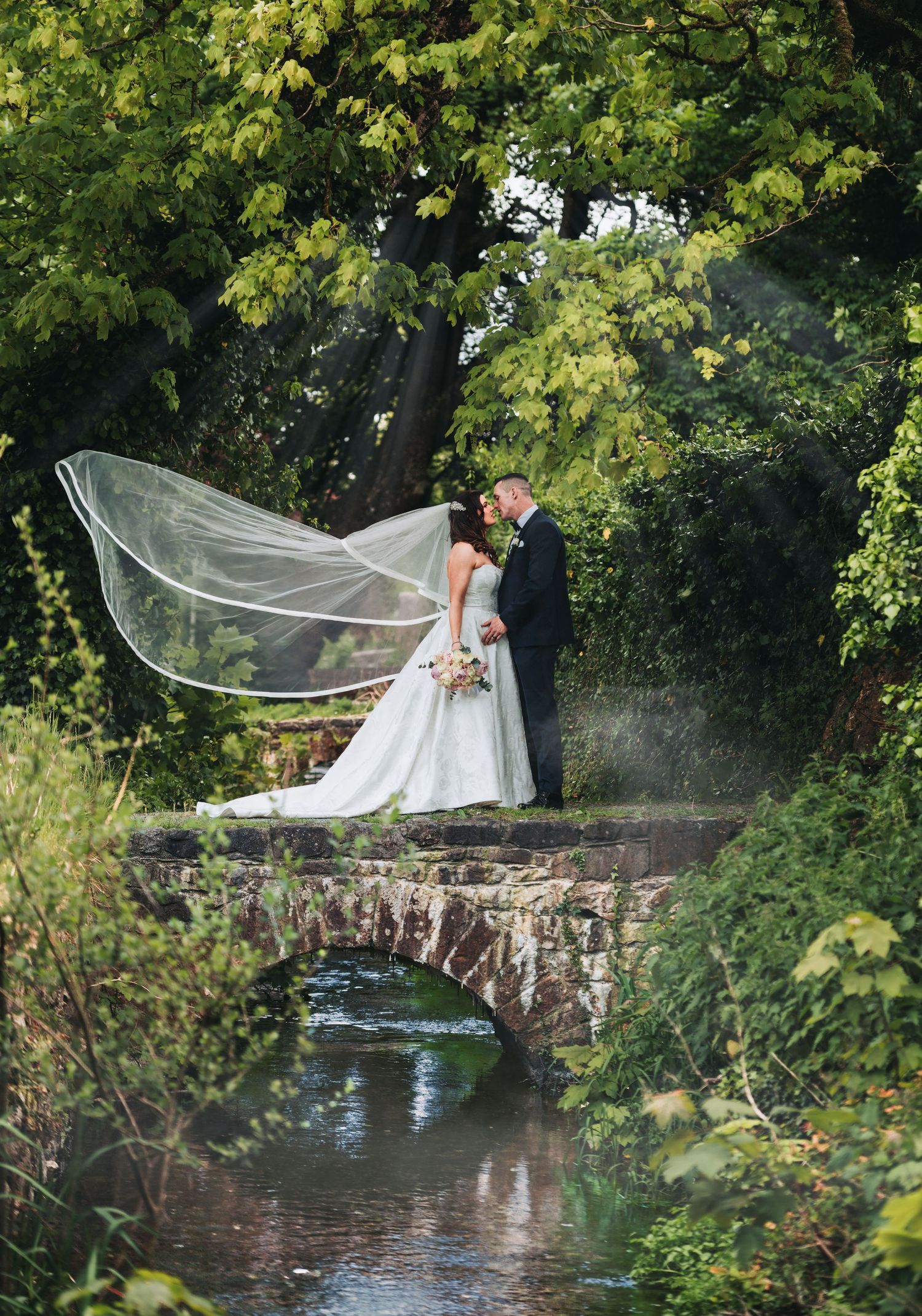 Bride and groom kissing on a stone bridge with veil flowing in the wind, surrounded by lush greenery.