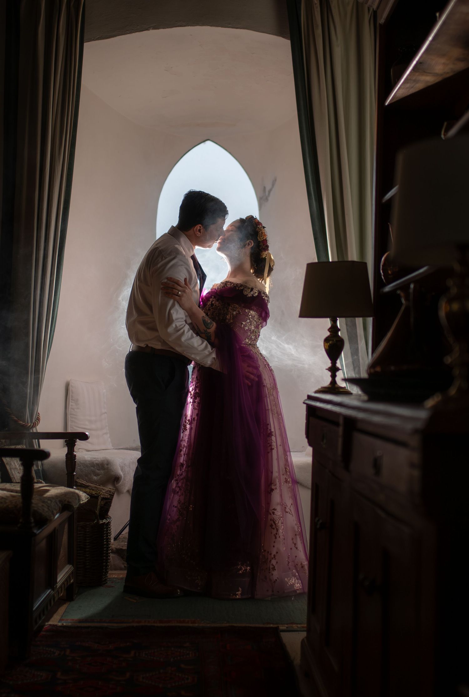 Couple sharing a romantic kiss in a dimly lit room with an arched window, woman in elegant burgundy gown.