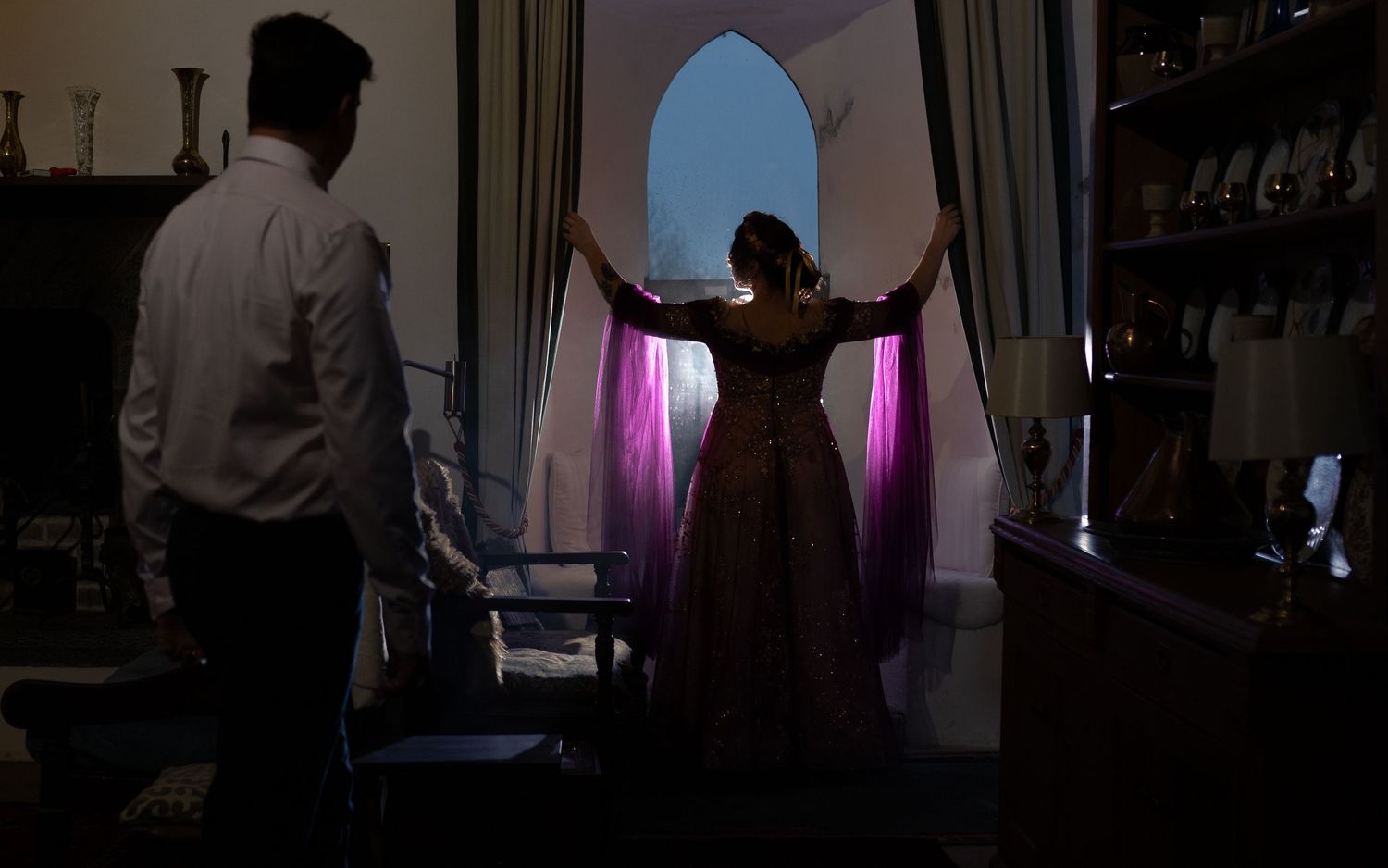 A woman in a elegant gown stands silhouetted by a glowing arched window with purple curtains in a dimly lit room.