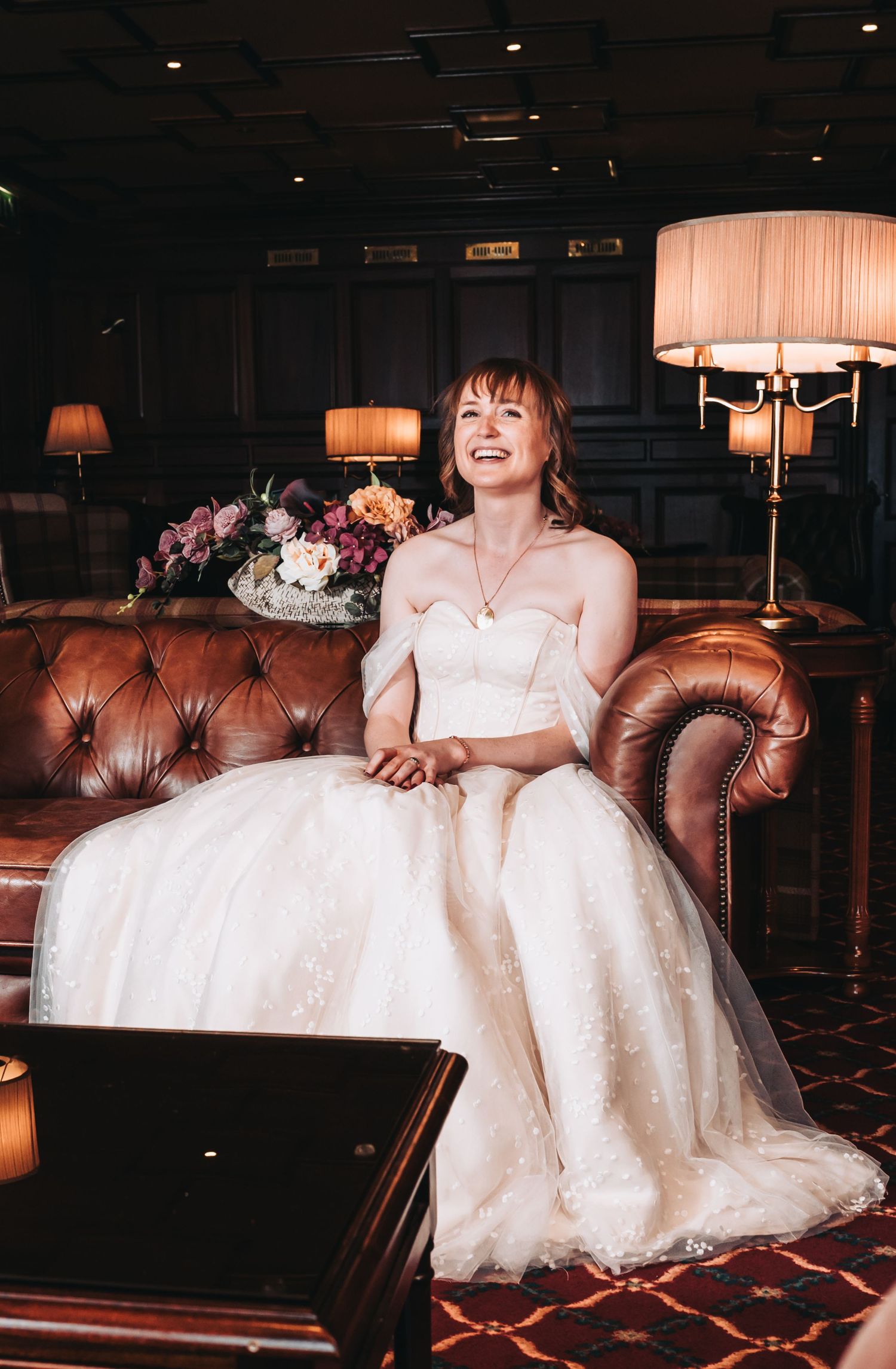 Smiling bride in strapless lace wedding gown seated on leather sofa surrounded by floral arrangements.