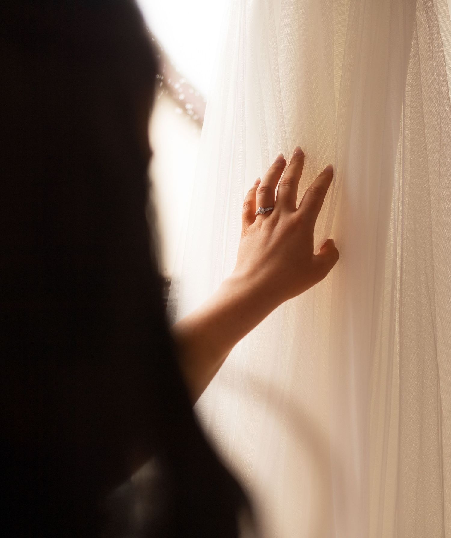A bride's hand with an engagement ring gently touches a flowing white wedding veil in soft, dramatic lighting.