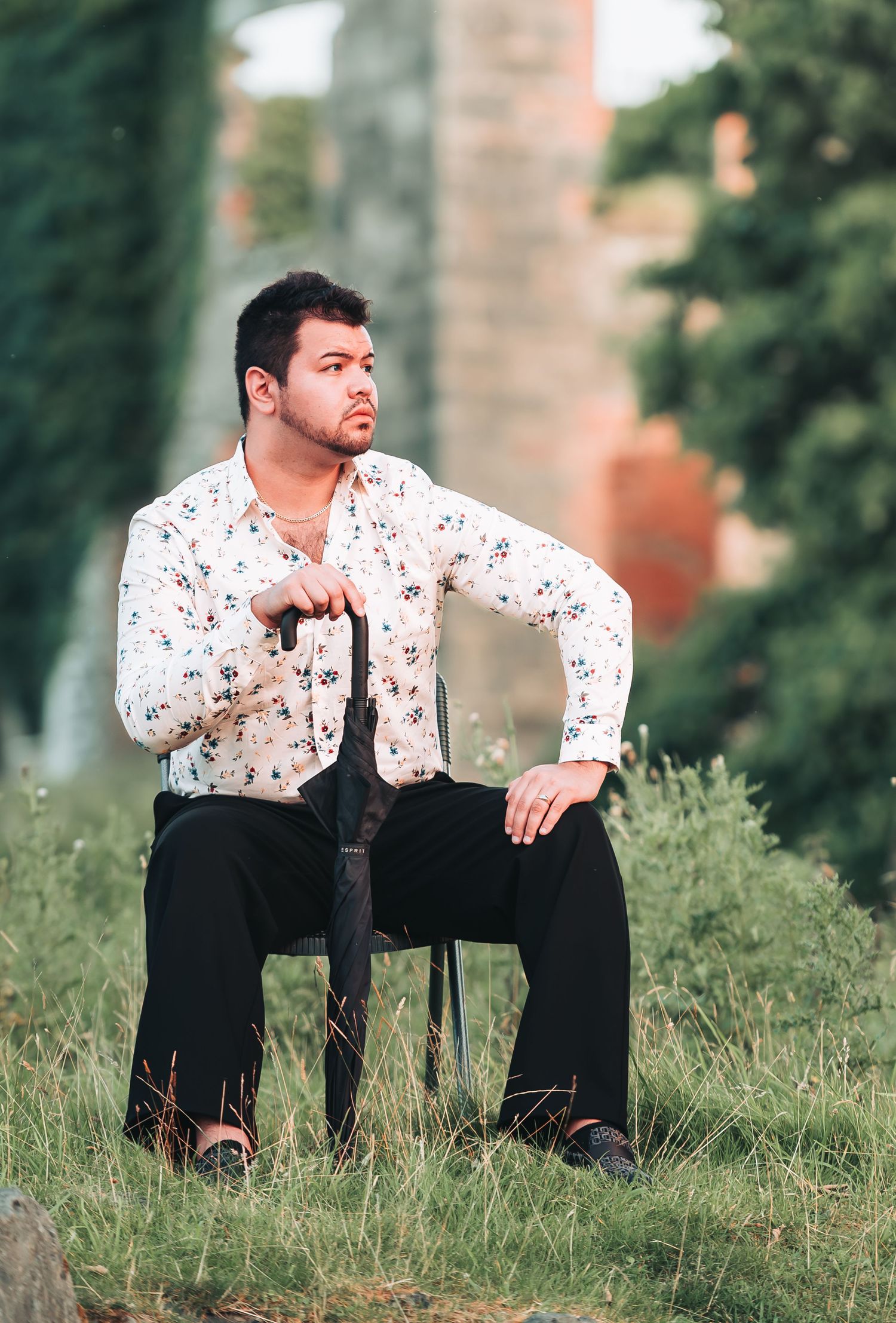 A man in a floral shirt sits on a chair outdoors, holding an umbrella, with a blurred green background.
