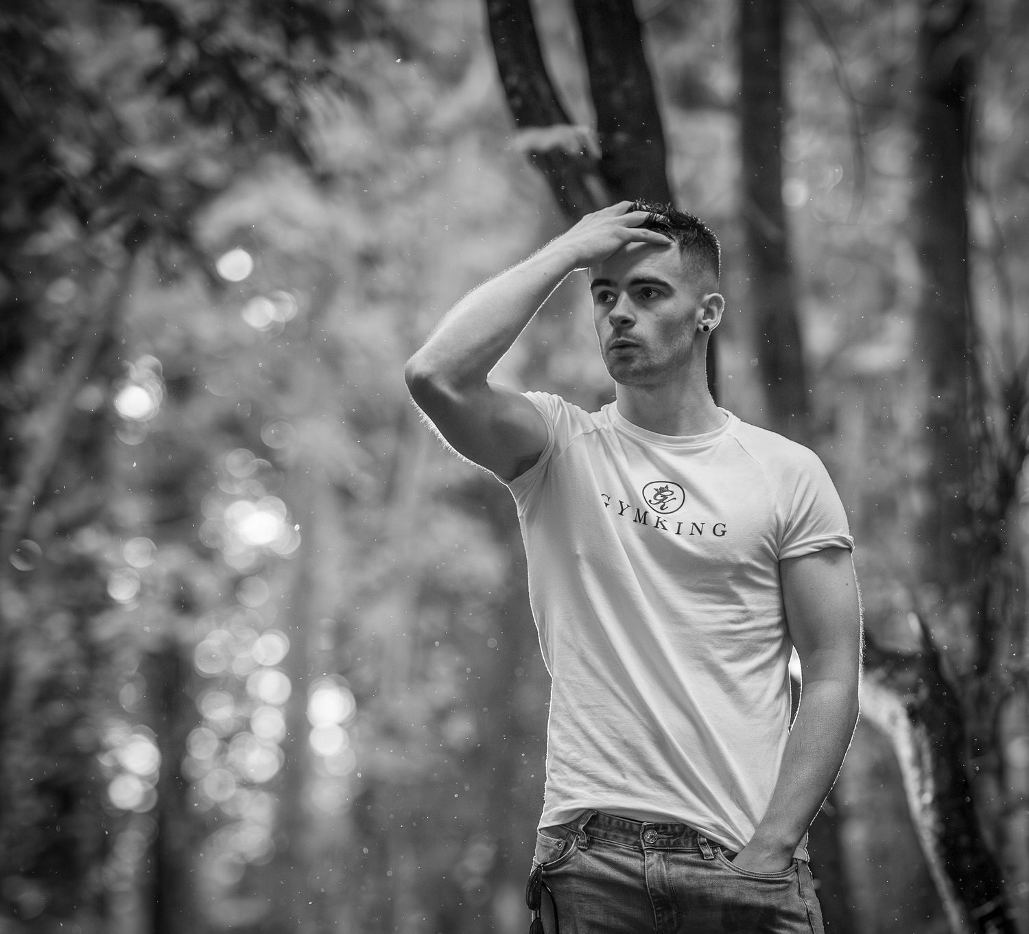 Black and white photo of a young man in a white t-shirt posing in a forest, hand raised to his forehead.