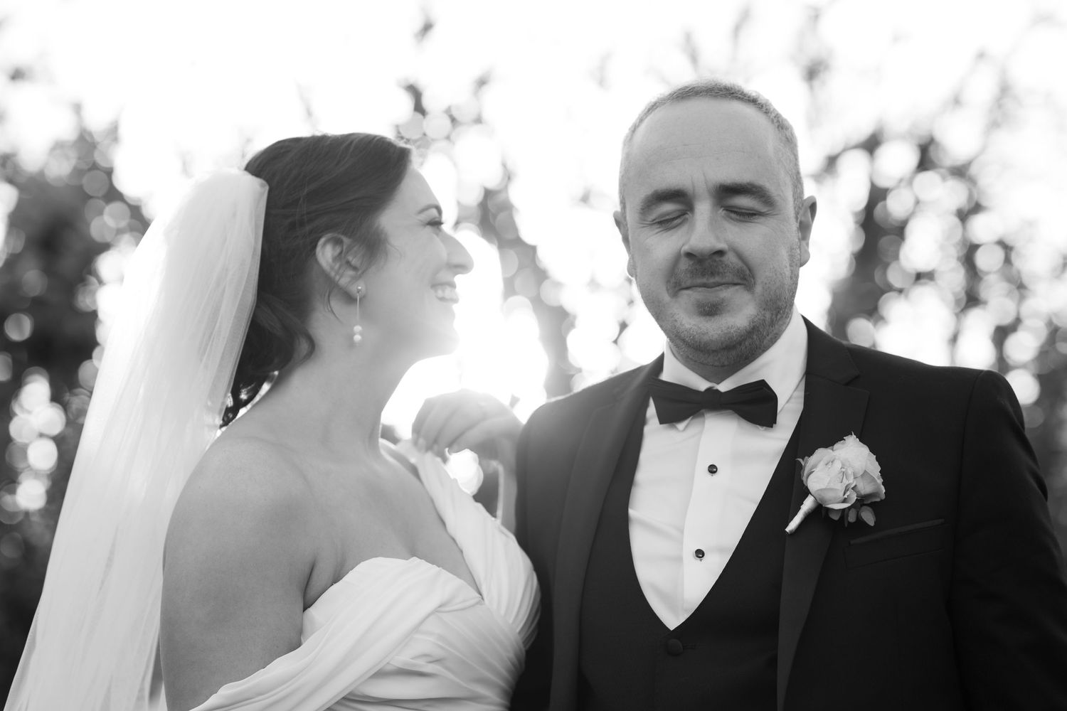 Black and white wedding portrait of bride in veil and groom in tuxedo with boutonniere smiling outdoors.